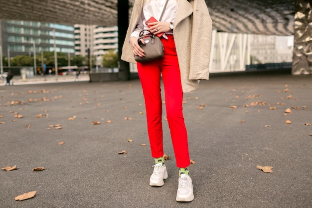 Close up fashion details , young woman wearing trendy red pants funny socks and ugly fashion sneakers, beige elegant coat, posing on the street near business centers, autumn time.