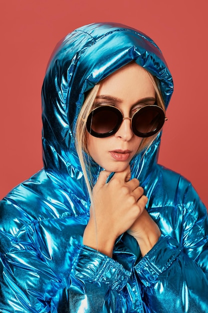 Close up fashion photo of young woman covering herself with hood. She wears blue oversized down jacket in studio on red background