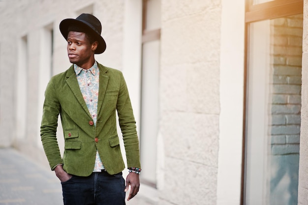 Close up fashion portrait of black african american man on green velvet jacket and black hat walking on streets of city background house with many windows