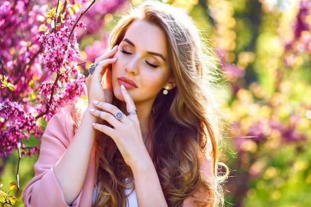 Close up fashion portrait of tender elegant pretty woman with big green yes and full lips, natural fresh make up and long fluffy hairs, paint neat Sakura blooming tree, sunny spring time.