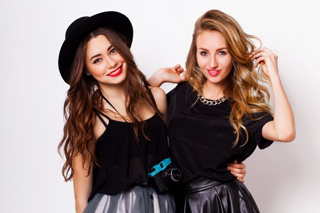 Close up fashion portrait of two elegant stylish  women wearing a leather  skirt and black hat , holding  retro camera.  Posing against white background.