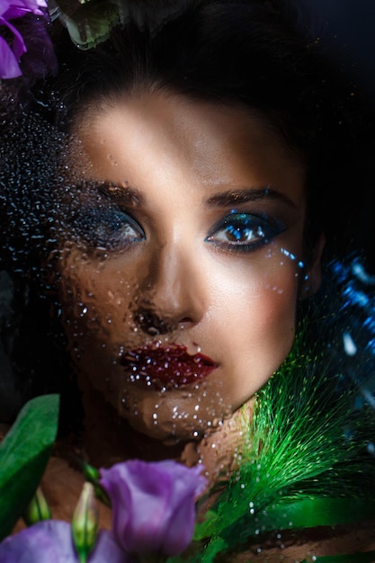 Close up fashion portrait of young beautiful brunette woman with bright make up looking at front through misted glass, lying among white and purple eustomas.