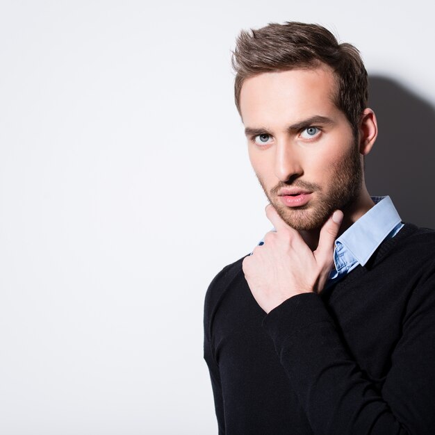 Close-up fashion portrait of young man in black pullover poses over wall with contrast shadows