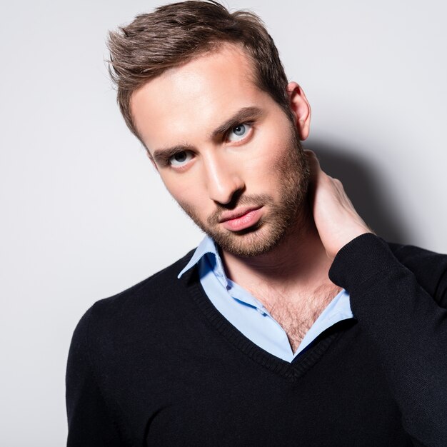 Close-up fashion portrait of young man in black pullover poses over wall with contrast shadows