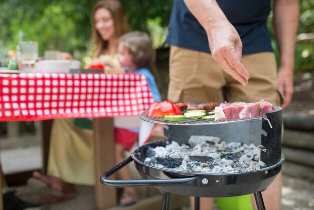 Close-up of father cooking BBQ. Mid adult man standing at barbeque grid, cooking fresh meat. Mother and sons sitting at table together