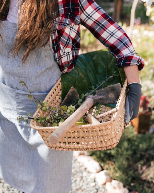 Primo piano di un giardiniere femminile che tiene zappa e ramoscelli raccolti nel cestino