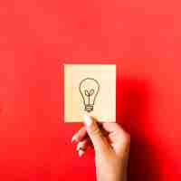 Free photo close-up of female hand holding sticky note with drawn light bulb on red background