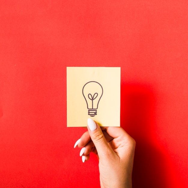 Close-up of female hand holding sticky note with drawn light bulb on red background