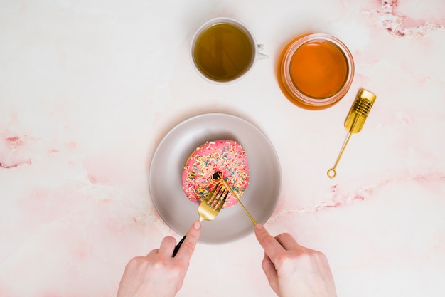 Close-up of a female hand's cutting the pink donut with fork and butter knife against textured backdrop