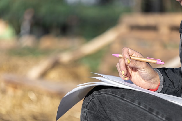 Foto gratuita il primo piano di una mano femminile con una manicure gialla tiene una penna e fogli di carta su uno sfondo sfocato all'esterno, copia spazio.