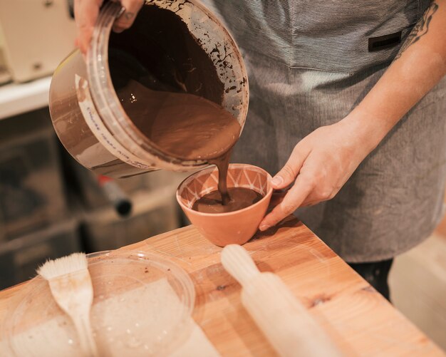 Close-up of female potter's hand pouring the brown paint in the bowl on wooden table