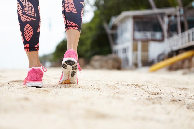 Close up of female runner wearing pink sneakers and leggings walking or running on beach sand while exercising outdoors against blurred bungalow. View from the back.