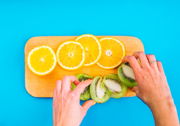 Foto gratuita primo piano della mano di una femmina che organizza le fette di kiwi con le arance sul tagliere contro fondo blu