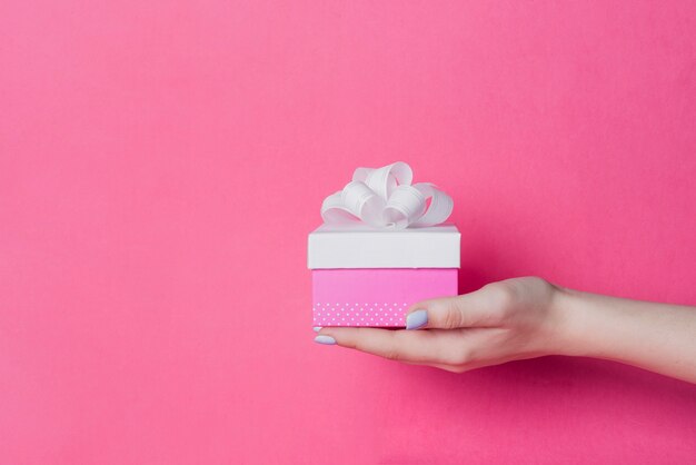 Close-up of female's hand holding box with white ribbon bow on pink background