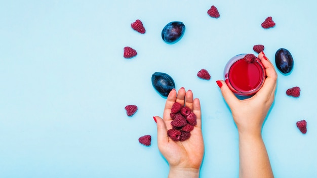 Close-up of female's hand holding raspberries and juice with plums on blue background