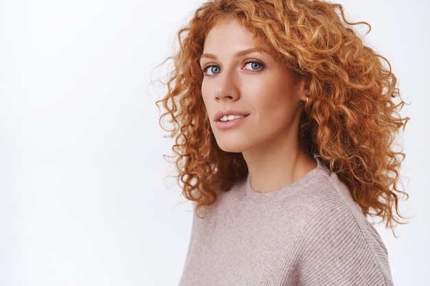 Primo piano femminile splendida rossa donna riccia in camicetta beige in piedi mezzo capovolta sul muro bianco, gira la testa verso la telecamera con un'espressione sensuale, felice e civettuola, flirtando