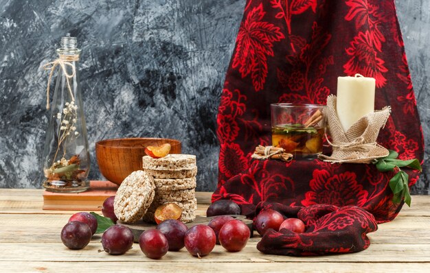 Close-up fermented drink and candle on red scarf with wafers,jug vase,a bowl,plums and red scarf on wooden board and dark grey marble background. horizontal free space for your text