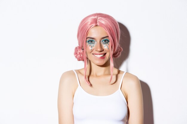 Close-up of flirty attractive female model in pink wig, with bright party makeup, biting lip and smiling, looking at something tempting, standing.