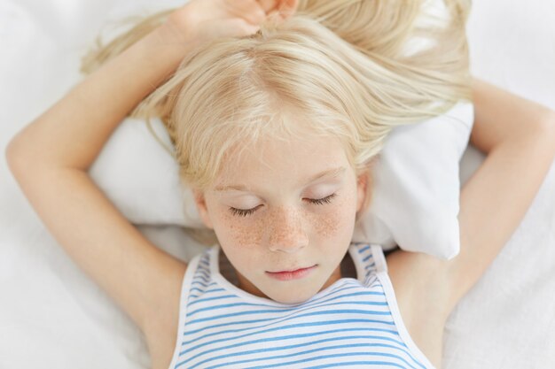 Close up of freckled girl with blonde hair lying on white bedclothes, sleeping at night and having pleasant dreams. Small female child daydreaming. Realxed female kid closing eyes, feeling relaxation