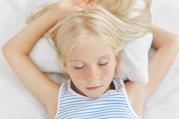 Close up of freckled girl with blonde hair lying on white bedclothes, sleeping at night and having pleasant dreams. Small female child daydreaming. Realxed female kid closing eyes, feeling relaxation