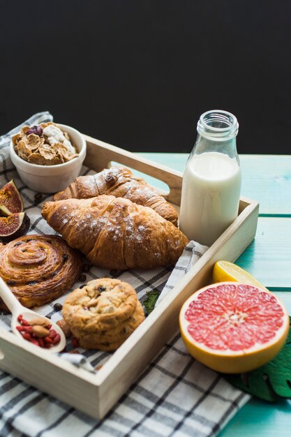 Close-up of fresh croissant; backed cookies; milk; muesli; and citrus fruit with cloth in wooden container