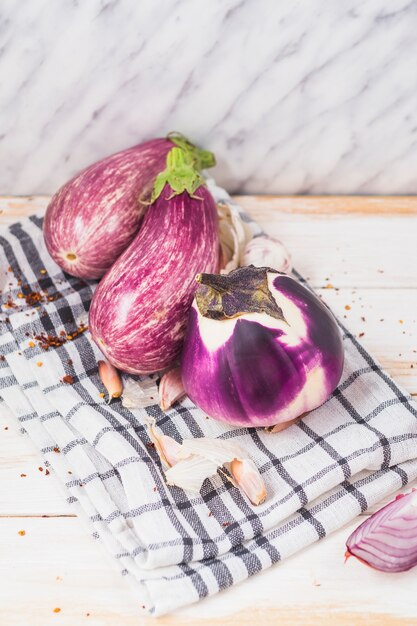 Close-up of fresh eggplants; onion; garlic cloves; chili flake and cloth on wooden tabletop