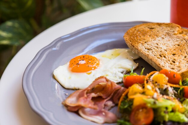 Close-up of fresh fried egg; bacon; toast and salad served on gray plate over the white table