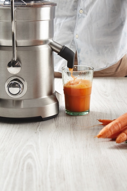 Free photo close up fresh juice pours from steel professional juicer to transparent glass in cafe shop. healthy drink for breakfast from fresh organic carrots, lying on wooden table near