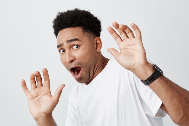 Close up of funny attractive dark-skinned african male student with curly hair in casual white t-shirt spreading hands with frightened expression, watching scary movie in cinema.