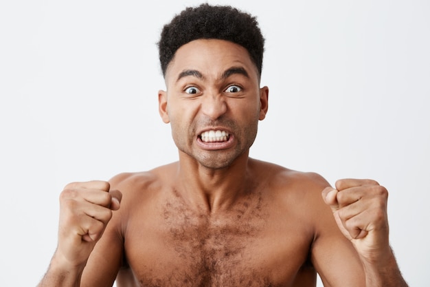 Close up of funny attractive dark-skinned african male with curly hair looking TV with mad expression, cheering for his favorite baseball team, screaming and gesticulating with hands.