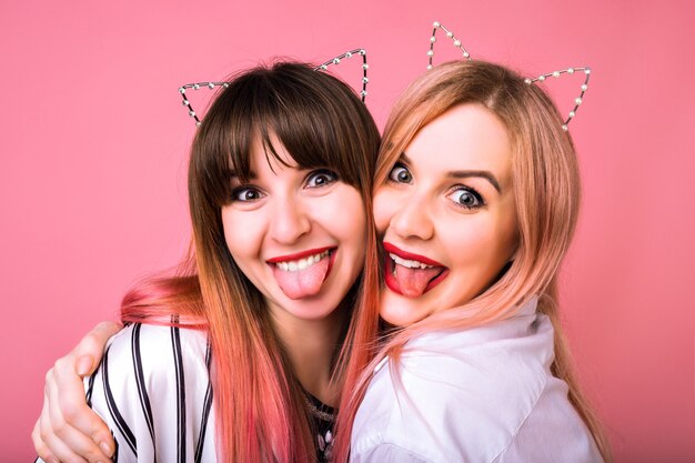 Close up funny crazy portrait of happy girls having fun showing tongues and wearing party cat ears, pink wall and youth style of hairs, best friends hugs.