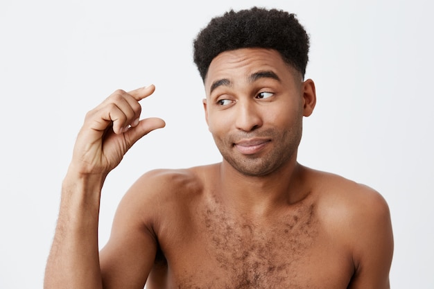 Close up of funny dark-skinned male with afro hairstyle and naked body showing little sign with hand, looking aside with sarcastic expression. People's emotions.