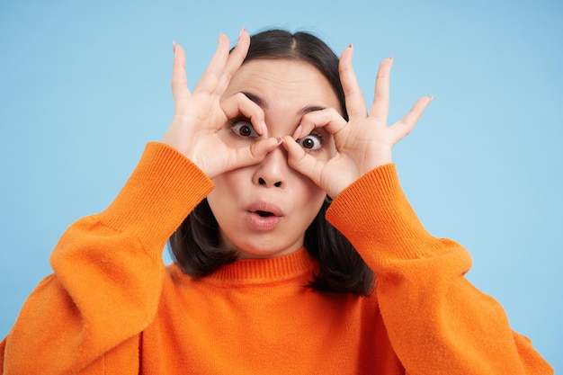 Close up of funny korean girl shows glasses and makes silly face stands over blue background