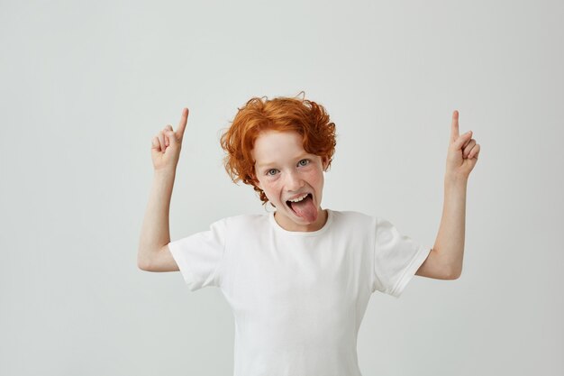 Close up of funny little boy with curly ginger hair and freckles pointing up with both hands, having silly face with open mouth. Copy space.