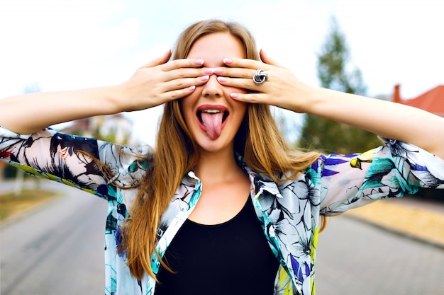 Close up funny portrait of smiling blonde girl close her eyes buy her hands, bright shirt, countryside, shoeing her long tongue, bright manicure.