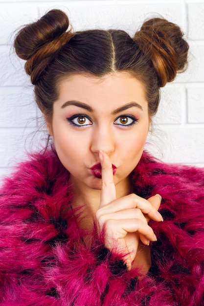 Close up funny portrait of young stylish sexy girl with cool hairstyle and bright trendy make up, wearing stylish fake fur coat, eighties style. Put her finger to the lips and say shhh