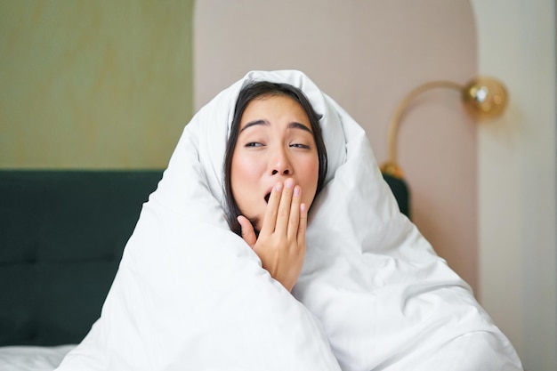 Free photo close up of funny sleepy girl asian woman yawns after waking up early morning covers herself with bl