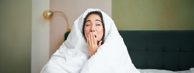 Foto gratuita primo piano di una donna asiatica divertente e assonnata che sbadiglia dopo essersi svegliata la mattina presto e si copre