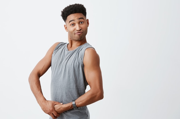 Close up of funny young afro athlete with curly dark hair playing with muscles, looking in camera with funny face expression. Health and beauty. Copy space