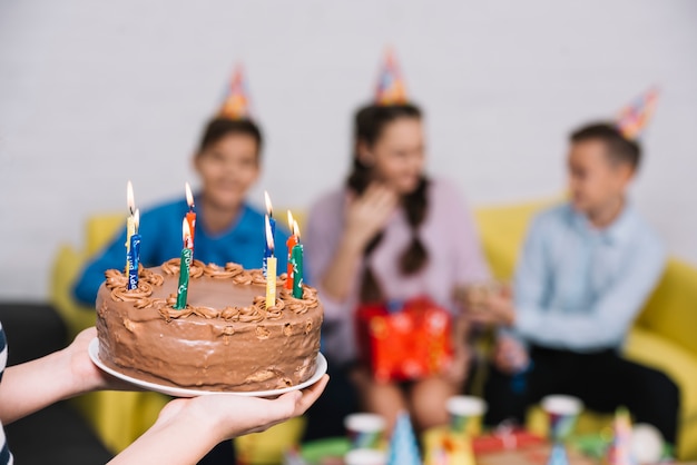 Close-up of a girl bringing chocolate cake decorated with lighted candles to her friends