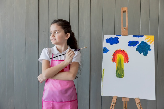 Close-up of a girl holding paint brush in hand standing near the easel looking away