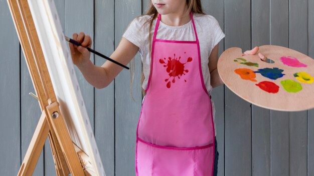 Close-up of a girl holding palette in hand painting on the easel with paint brush