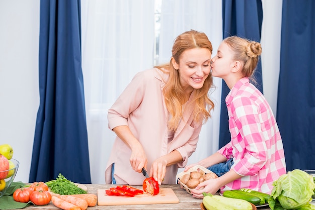 Foto gratuita primo piano di una ragazza che bacia sua madre che taglia le verdure con il coltello nella cucina