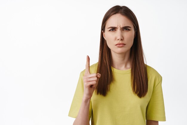 Close up of girl makes confused face, dont get it, pointing up, frowning and squinting puzzled, cant understand smth, standing on white.