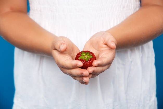 Close up of girl's hand holding strawberry over blue wall