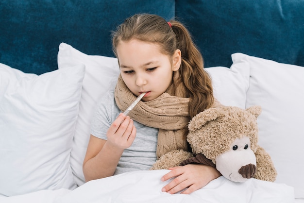 Close-up of a girl suffering from the cold inserting the thermometer in her mouth sitting on bed with soft toy