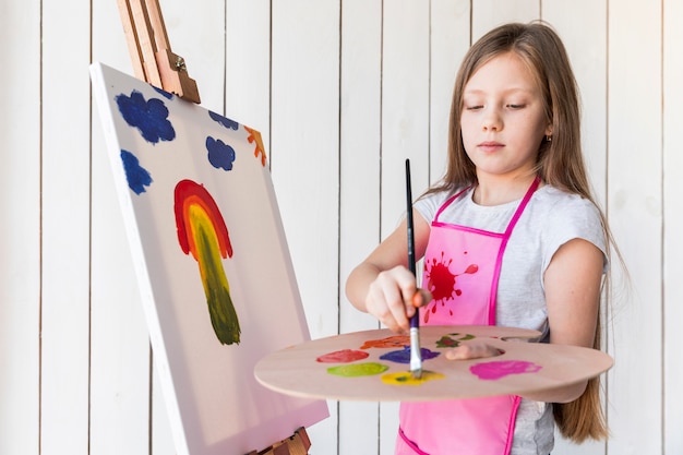 Free photo close-up of a girl with long blonde hair painting on the canvas standing against white wooden wall