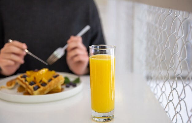 Close-up of a glass of orange juice on a blurred background, a woman having breakfast with Belgian waffles and juice.