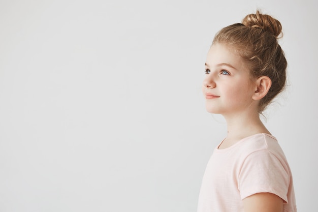 Close up of good-looking little girl with blonde hair in bun hairstyle, standing in three quarters, looking aside with smile on her face.