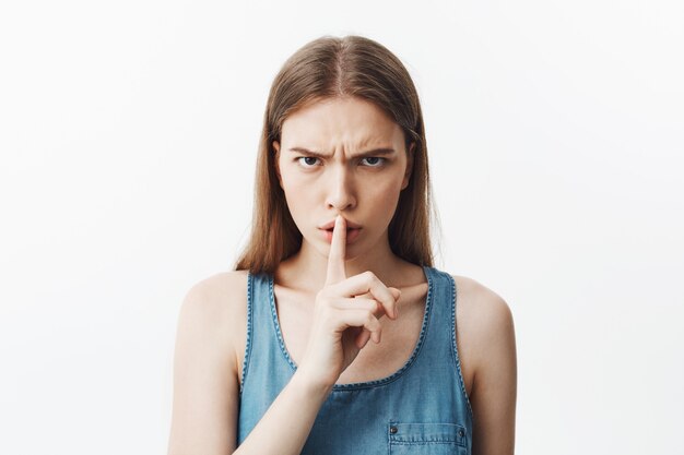 Close up of good-looking young caucasianstudent girl with dark hair in blue shirt holding hand in fromt of lips, making hush gesture with serious and unsitisfied expression. Woman tries to calm down l